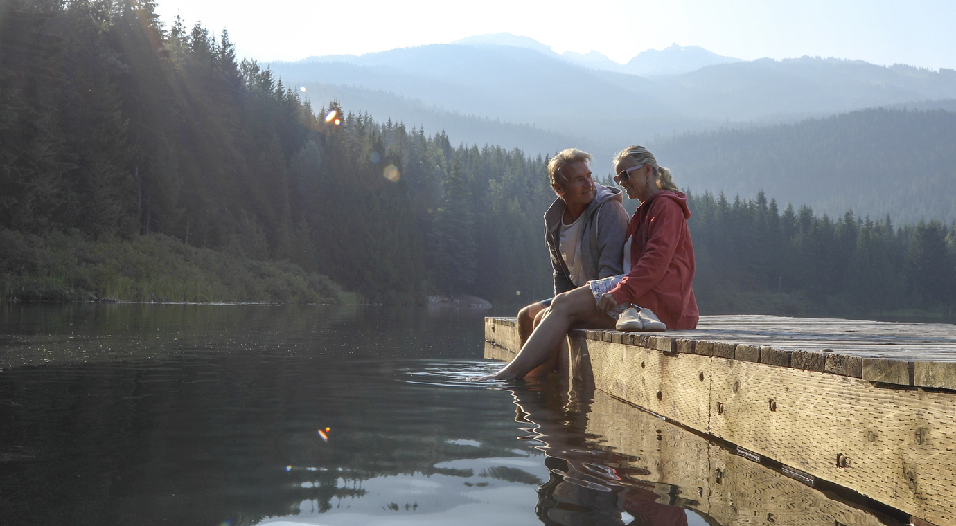 Mature couple relax on wooden pier, looks out across lake Living in Canada